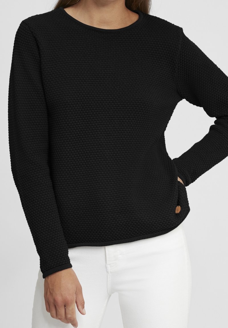 Femme portant un pull noir texturé à manches longues avec un col rond et un pantalon blanc, main posée sur la hanche devant un fond uni.
