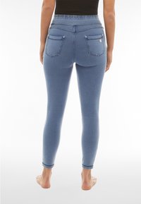 Nainen, jolla on vaaleansiniset kapeat farkut ja musta yläosa, seisoo paljain jaloin valkoista taustaa vasten, kuvattuna takaa.