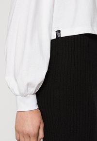 Weißes Langarmoberteil mit voluminösen Ärmeln, ribbelige schwarze Hose und ein kleines schwarzes "Positive"-Etikett am Saum des Oberteils.