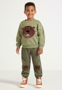 Felpa verde oliva con design di un volto di animale in peluche. I pantaloni a coste coordinati presentano toppe di animali simili sulle ginocchia. Scarpe alte arancioni.