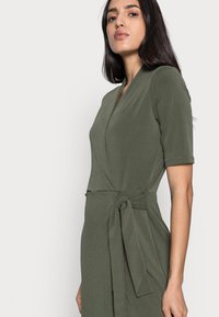 Vestido envolvente verde-oliva com mangas curtas, apresentando um detalhe de laço na cintura e uma textura de tecido suave.