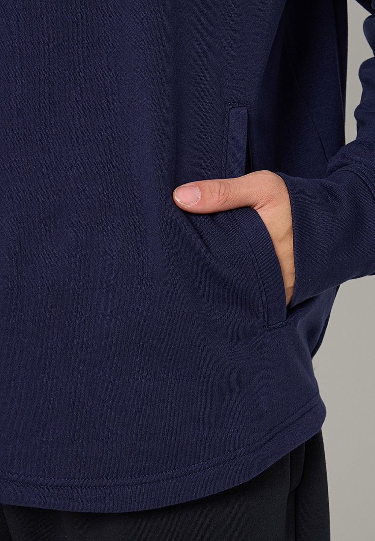 Mano colocada dentro del bolsillo de un sudadera azul marino, mostrando la textura de la tela y los detalles de la costura en la prenda.