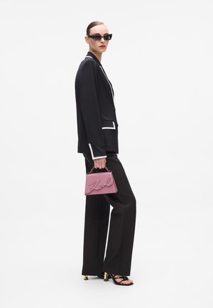 Black blazer with white trim, black trousers, black sunglasses, pink handbag with "Karl" in cursive, and strappy black sandals.
