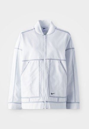 Hvit Nike-jakke med glidelås foran, høy krage, fremtredende sømmer, to frontlommer og en liten svart logo nederst foran.