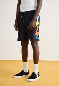 Shorts de bain noirs avec cordon de serrage, ornés de graphiques de foudres colorées en orange, jaune et aqua. Portés avec des chaussures noires à enfiler et des chaussettes blanches.