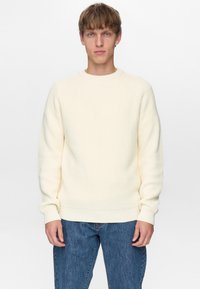Cremefarvet strikket sweater med ribstrikket tekstur, rund halsudskæring og raglanærmer, kombineret med blå denimjeans.