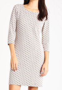 Robe beige clair avec un motif géométrique blanc et marron, décolleté rond, manches trois-quarts et coupe ajustée. Texture de tissu douce.