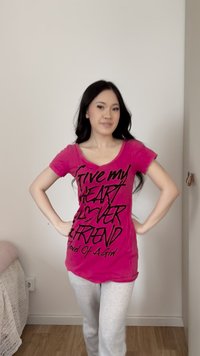 Ung kvinne med langt mørkt hår står med hendene på hoftene, iført en knallrosa skjorte med svart tekst og lysegrå bukser i et rom.