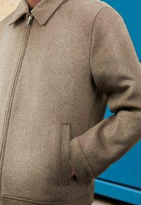 Braune Wollmischungjacke mit Reißverschluss, spitzem Kragen und Seitentaschen. Strukturierter Stoff und lockerer Schnitt mit klaren Linien.