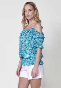 Top turchese a spalle scoperte con bordi arricciati, decorato con motivi floreali e geometrici bianchi. Abbinato a pantaloni corti bianchi.