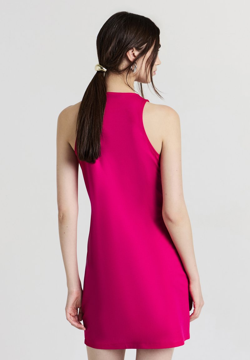 Donna con lungo capelli scuri raccolti in una coda di cavallo, che indossa un vestito senza maniche di un brillante colore rosa, rivolta di spalle contro uno sfondo chiaro e semplice.