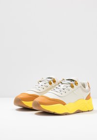 Sneakers featuring a white upper, orange and yellow accents, textured sole, and metallic gold detailing on the laces.