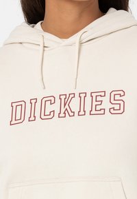 Krämfärgad hoodie tillverkad av mjukt tyg, med en framträdande mörkröd "DICKIES"-logotyp på framsidan. Justerbar snörning i huvan.