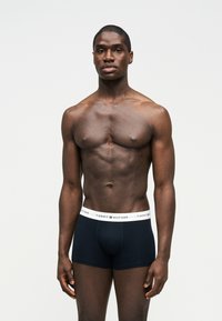 Μυώδης άνδρας που στέκεται μπροστά σε λευκό φόντο, φορώντας μπλε ναυτικό boxer Tommy Hilfiger με λευκή ζώνη μέσης με λογότυπο.