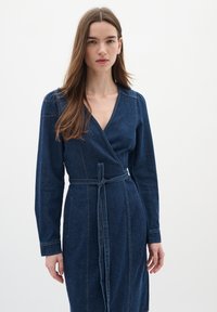 Denimomslagklänning i mörkblått med långa ärmar, v-ringning och midjebälte. Har sömdetaljer och en åtsittande design.