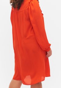 Robe à manches longues de couleur orange vif, confectionnée en tissu léger avec des fronces aux épaules et aux poignets, tombant au-dessus des genoux.