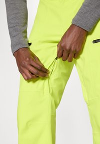 Χειρονομίες που ξεzipάρουν ένα πλαϊνό φερμουάρ σε φωτεινά κίτρινα παντελόνια, συνδυασμένα με ένα γκρι μπλουζάκι μακρύ μανίκι.