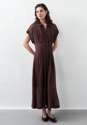 Frau mit dunklen, welligen Haaren trägt ein langes braunes Kleid mit kurzen Ärmeln und schwarze Ballerinas, steht mit den Händen hinter dem Rücken vor einer hellgrauen Wand.