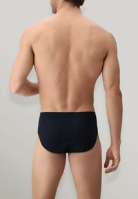 Slips da bagno neri realizzati in materiale liscio, con un taglio a vita bassa e una cintura elasticizzata. Posizionati su uno sfondo grigio chiaro.