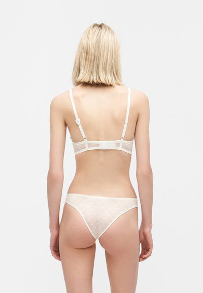Γυναίκα με κοντά ξανθά μαλλιά, φοράει διάφανο λευκό δαντελένιο σουτιέν και αντίστοιχο εσώρουχο, φωτογραφημένη από πίσω μπροστά σε απλό φωτεινό φόντο.