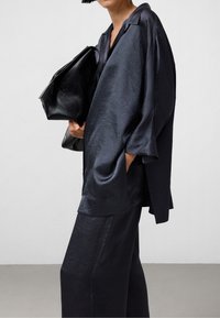 Blusa blu scuro dalla vestibilità ampia con una lucentezza texturizzata, dotata di colletto e maniche larghe, abbinata a pantaloni a gamba larga coordinati e una borsa nera.