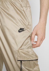 Beżowe spodnie Nike z czarnym logo i zapinaną na zamek kieszenią boczną, pokazane na prałym udzie osoby z widoczną ręką obok kieszeni.