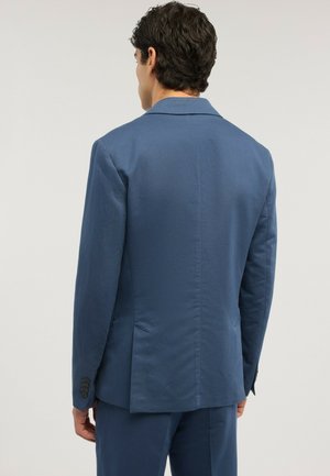 Mand med mørkt krøllet hår iført en tætsiddende blå jakkesætjakke og matchende bukser, set bagfra mod en ensfarvet lys baggrund.