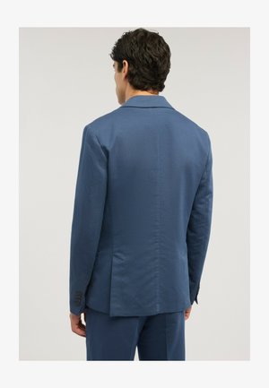 Mand med mørkt krøllet hår iført en tætsiddende blå jakkesætjakke og matchende bukser, set bagfra mod en ensfarvet lys baggrund.