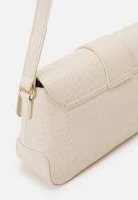 Světle béžová kabelka s embosovaným geometrickým vzorem, obdélníkového tvaru, bezpečným klopovým uzavřením a nastavitelným popruhem se zlatými kovovými detaily.