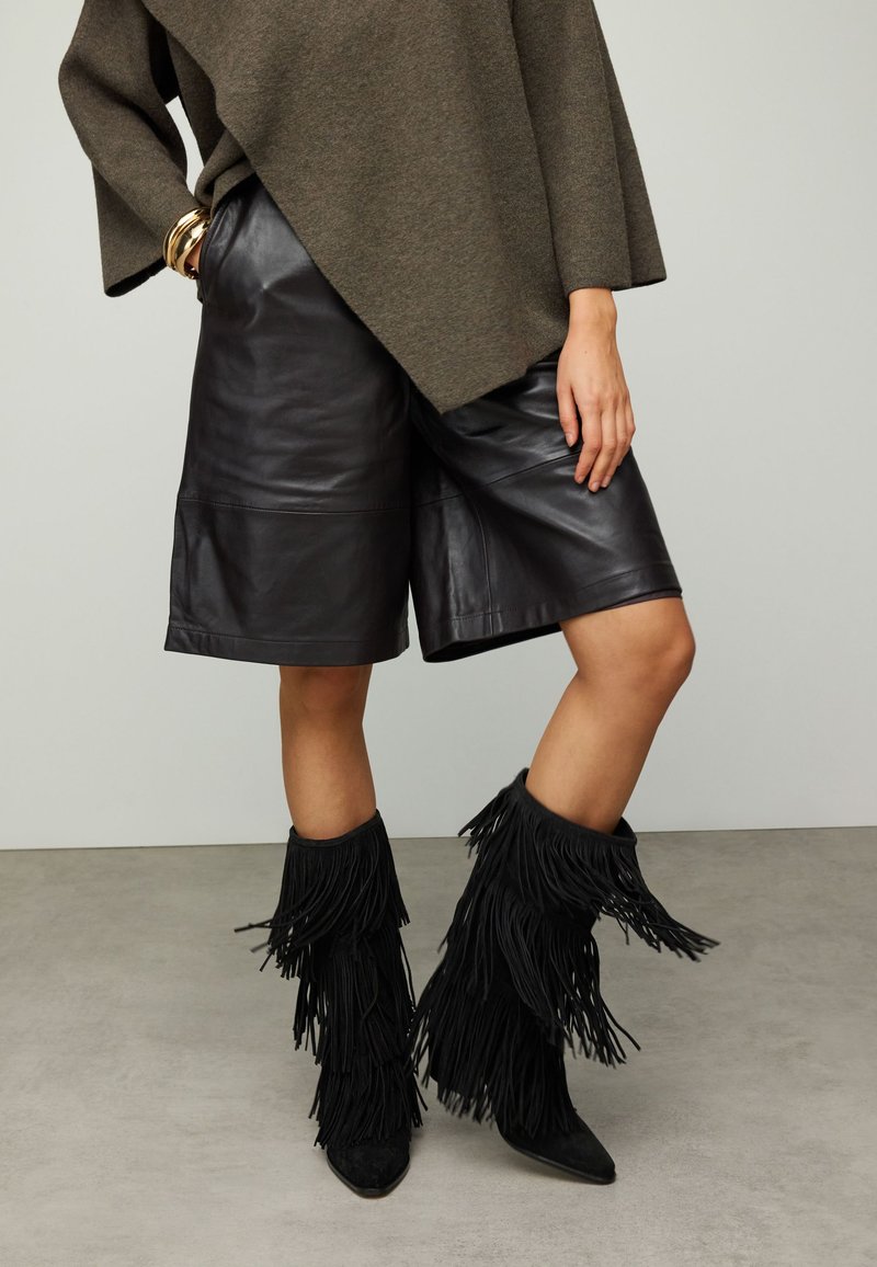 Shorts en cuir noir à coupe évasée, associés à des bottes hautes noires à franges et un pull oversize olive. Texture lisse, design en couches.