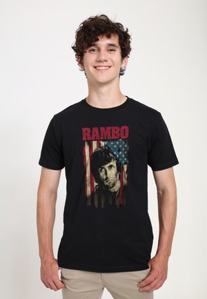 Young man with curly hair smiling, wearing a black t-shirt with a vintage Rambo graphic and American flag design.