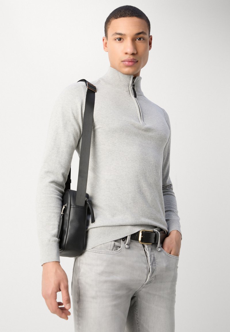 Maglione grigio chiaro con zip in un morbido filato, abbinato a jeans grigi chiari e una borsa a tracolla nera. Una cintura nera con una fibbia dorata completa l'outfit.
