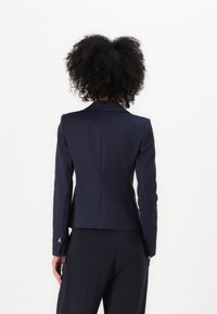 Marineblå skreddersydd blazer med strukturerte klaffer, tettsittende silhuett, glatt stoff og broderte detaljer på mansjettene. Bakvisningen viser rene linjer.