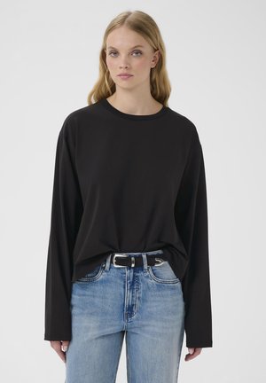 Mladá žena s dlhými blond vlasmi, ktorá má na sebe voľnú čiernu blúzu s dlhým rukávom zastrčenú do svetlomodrých džínsov s čiernym opaskom, stojaca neutrálne.