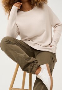 Beiger, gerippter Pullover mit langen Ärmeln, kombiniert mit olivgrünen Cargohosen und weißen Turnschuhen. Sitzt auf einem hölzernen Hocker.