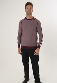 Suéter de punto burdeos con rayas blancas contrastantes en las mangas, cuello redondo y puños de canalé. Combinado con pantalones negros.