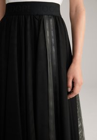 Jupe en tulle noir avec une large taille mettant en avant un logo en relief. Des rubans latéraux en gris foncé avec un motif de logo répété ajoutent du détail.