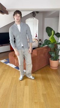 Gilet zippé en ribbed gris sur une chemise blanche, jeans gris clair et baskets argentées. Canapé en velours côtelé marron et plante en pot en arrière-plan.