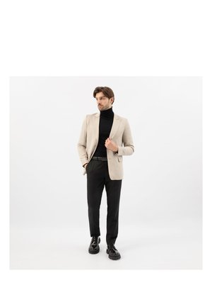 Man draagt een beige blazer, zwarte coltrui, zwarte broek en zwarte gepolijste schoenen, staande tegen een witte achtergrond, kijkt opzij.