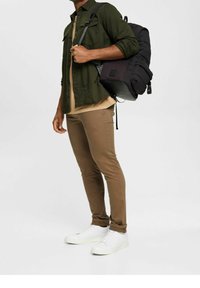 Zwarte rugzak op een lichtbruine shirt, olijfgroene jas en beige broek. Witte sneakers maken de outfit compleet. Minimalistische stijl en ontwerp.