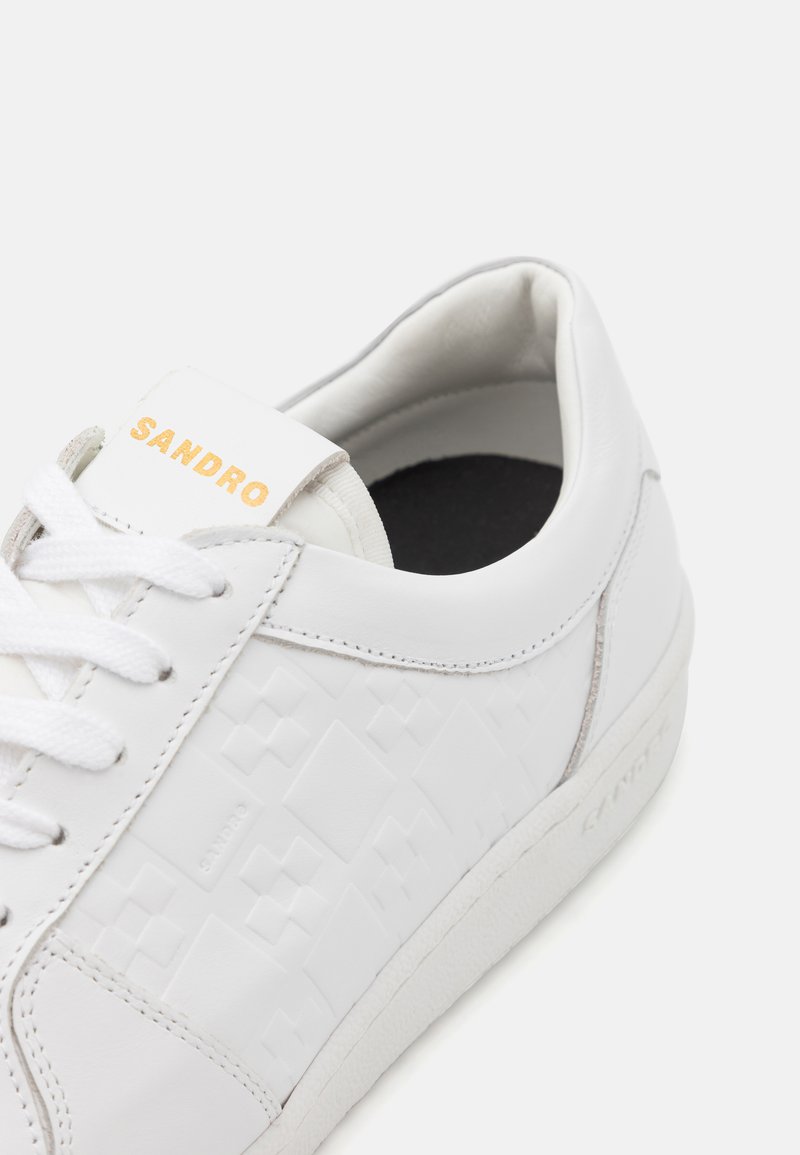 White leather sneaker with embossed geometric pattern, rounded shape, lace-up closure, and gold lettering on the tongue.