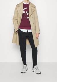 Trench coat beige de doble botonadura, sudadera burdeos con un logo gráfico, camisa blanca de botones, pantalones negros ajustados, zapatillas gris claro.
