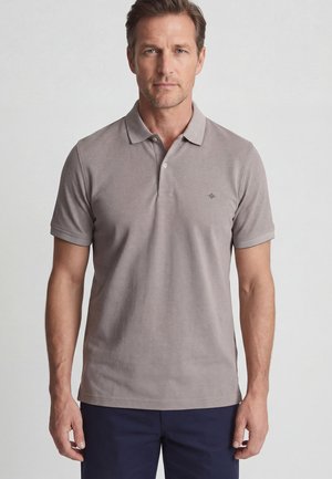 Man draagt een lichtgrijs polo met korte mouwen en een klein logo op de borst, staand tegen een effen witte achtergrond.