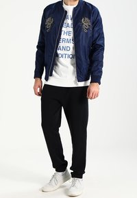 Marineblaues Bomberjacke mit Drachenstickerei, weißes Grafik-Sweatshirt, schwarze Jogginghose und weiße Sneaker, mit einem lässigen Schnitt.