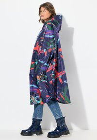 Poncho impermeabile sovradimensionato e colorato con una base blu navy, motivi astratti in rosso e verde, e un cappuccio. Abbinato a jeans blu e stivali neri lucidi.