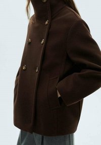 Cappotto doppiopetto marrone con colletto alto, caratterizzato da bottoni rotondi, tasche laterali e una silhouette strutturata. Realizzato in tessuto morbido.