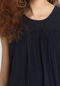 Vestido sin mangas azul marino con un diseño texturizado y fluido; presenta un cuello redondo y ligeros fruncidos en el busto. Pendientes grandes de aro visibles.