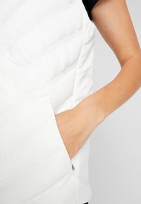 White puffer vest with quilted pattern, side pocket, and slight sheen. Smooth texture with a round neckline and sleeveless design.