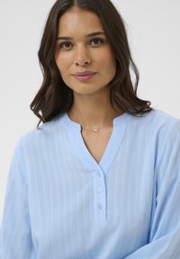 Junge Frau mit schulterlangem, braunem Haar, die eine hellblaue, geknöpfte Bluse und eine filigrane silberne Halskette trägt, vor neutralem Hintergrund.