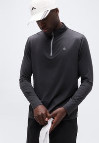 Άνδρας που φοράει σκούρα γκρι μακρυμάνικο πουκάμισο με φερμουάρ και λευκή καπέλο, κρατώντας ένα λευκό ύφασμα με το κεφάλι ελαφρώς σκυμμένο σε απλό φόντο.